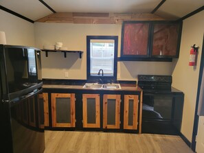 Een compacte keuken met houten kastjes, een zwart fornuis, een spoelbak en een raam met zonwering.