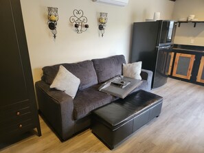 Een moderne woonkamer met een donkerbruine bank, een passende voetenbankje en een klein bijzettafeltje. Er zijn decoratieve wandlampen en een zwarte koelkast op de achtergrond.
