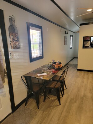 Een eetruimte met een lange tafel, vier stoelen, een tafel met glazen blad en een raam met jaloezieën.