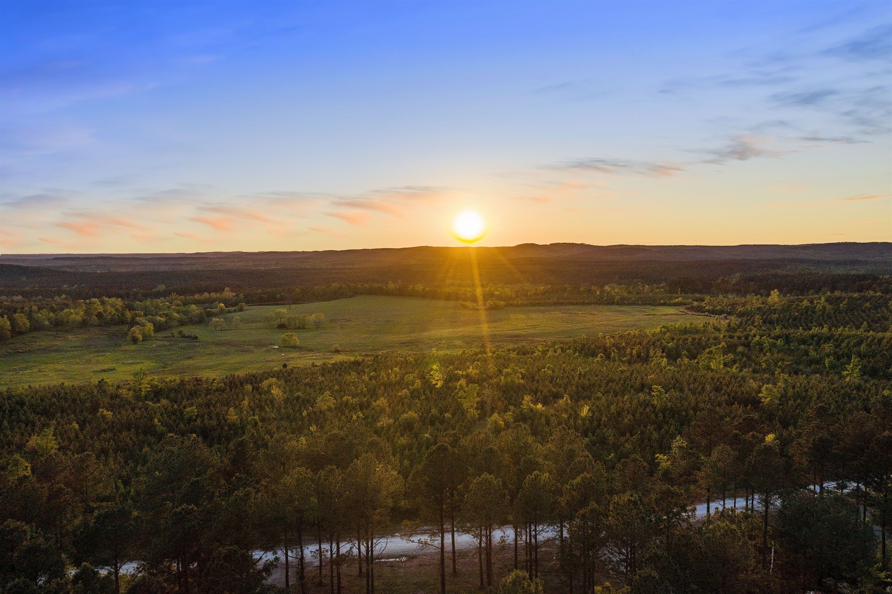 Laaja panoraamanäkymä metsäisestä maisemasta, jossa aurinko laskee kukkuloiden taakse.