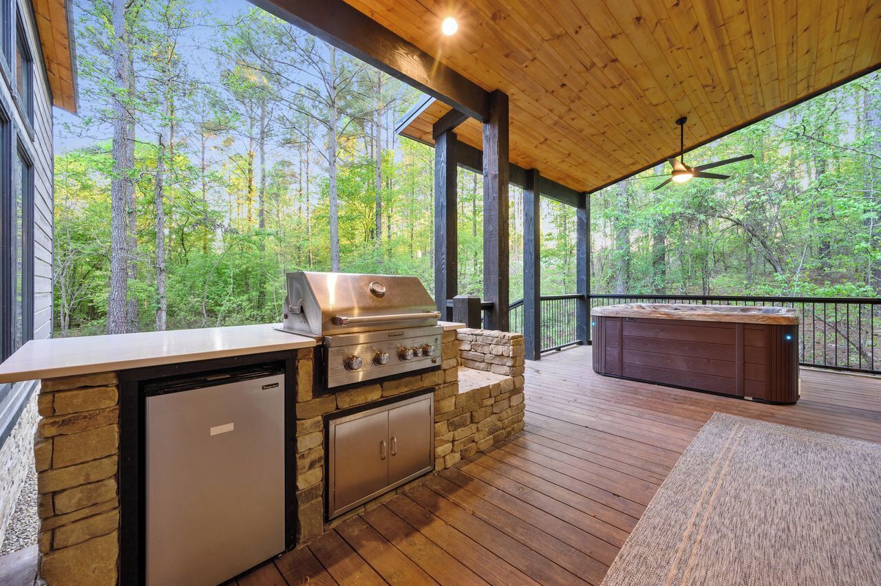 Una cocina al aire libre con una parrilla, un refrigerador empotrado y un jacuzzi en una terraza de madera con vistas a un bosque.