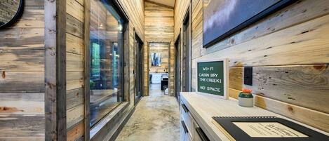 Un pasillo estrecho con paredes de madera y ventanas amplias, que conduce a una zona de cocina. Un cartel en la pared dice 'CABIN BRY THE CREEK NO SPACE'.