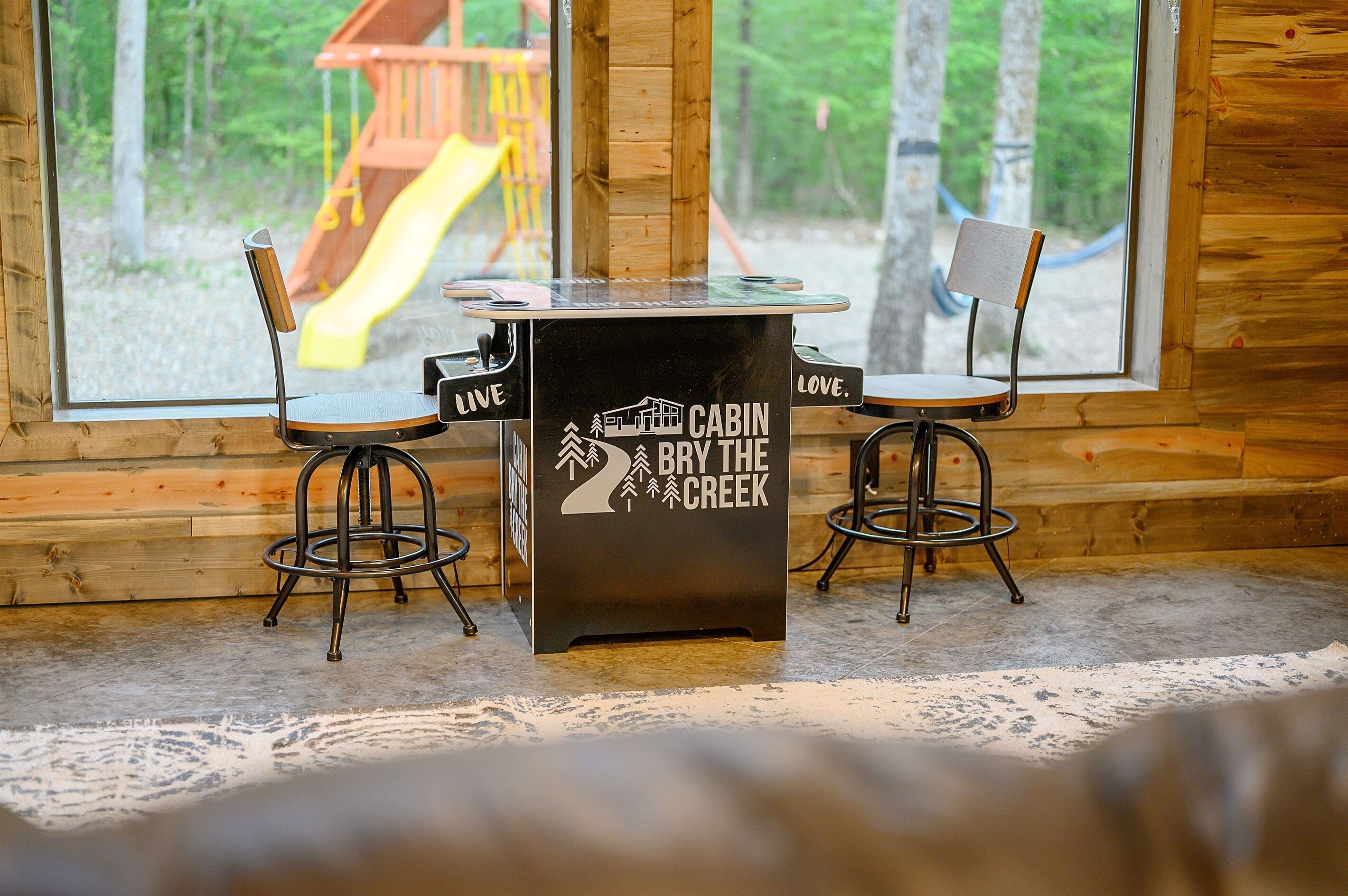 A cabin interior with a table and two chairs, a window with a view of a playground, and a wooden floor.