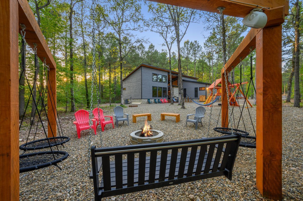 A wooden swing set, a fire pit, and a building with a deck in a forested area.