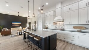 Une cuisine moderne avec des armoires blanches, un grand îlot central doté de tabourets, des luminaires suspendus et un dosseret en carreaux disposés en motif chevron.