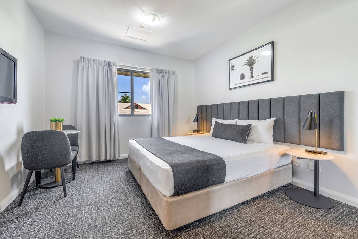 Habitación de hotel con cama, escritorio, silla, ventana con cortinas e imagen enmarcada en la pared.