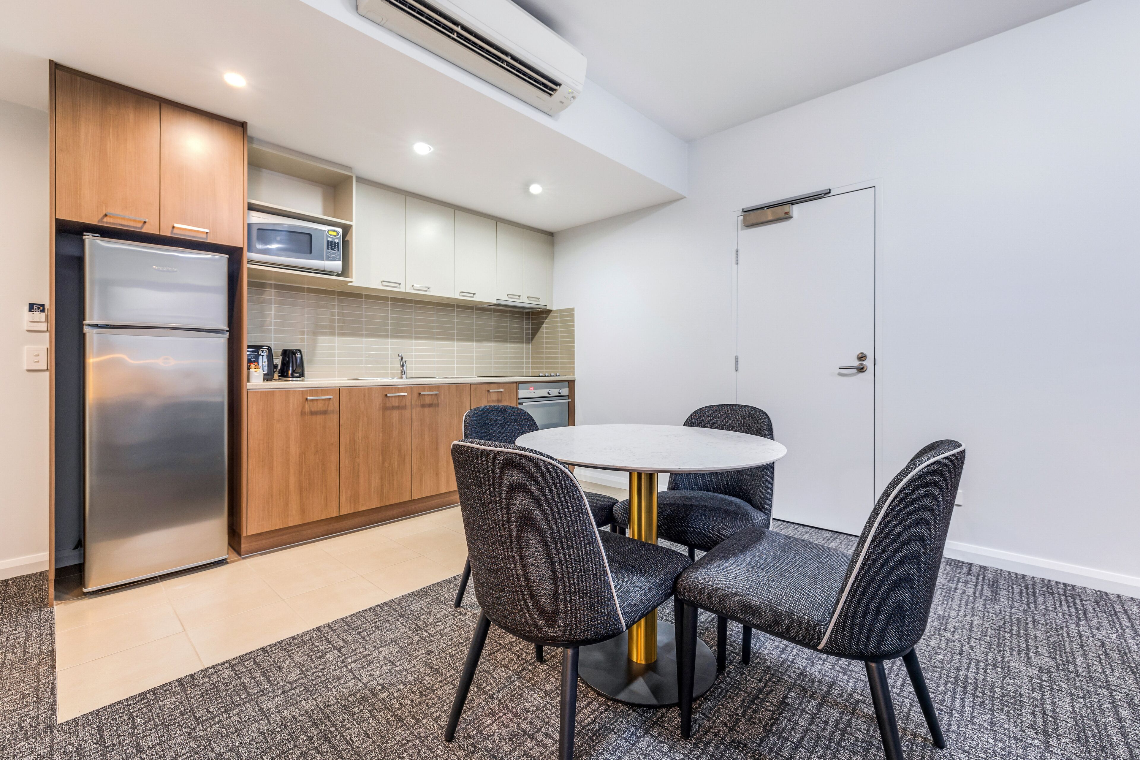 A compact kitchen with a refrigerator, microwave, and wooden cabinets. There is a dining table with four chairs and a carpeted floor.