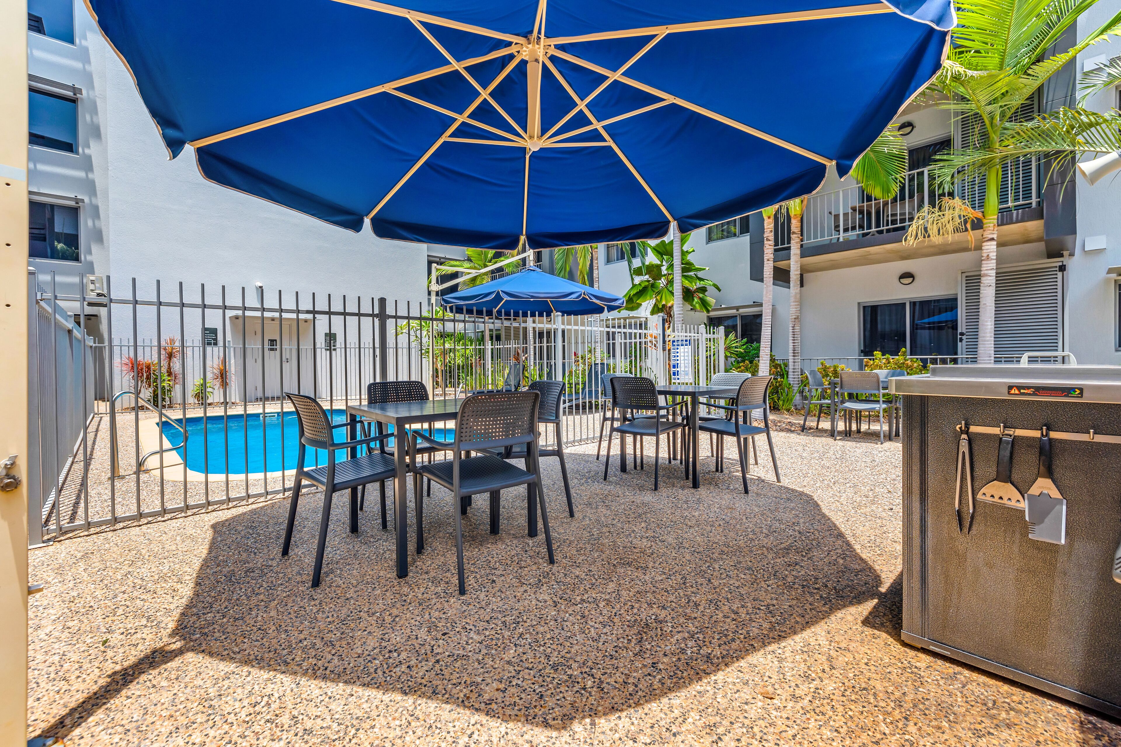 Un patio exterior con piscina, rodeado por una valla metálica, con mobiliario de exterior y una estación de barbacoa.