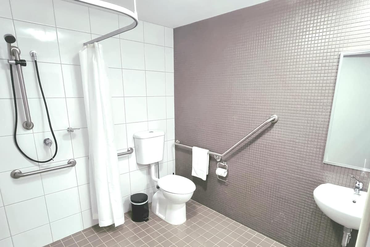 Un baño moderno con ducha, inodoro y lavamanos, equipado con barras de apoyo y un secador de manos.