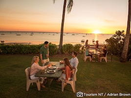 Un grupo de personas cenando al aire libre con vista al océano y a los barcos al atardecer.