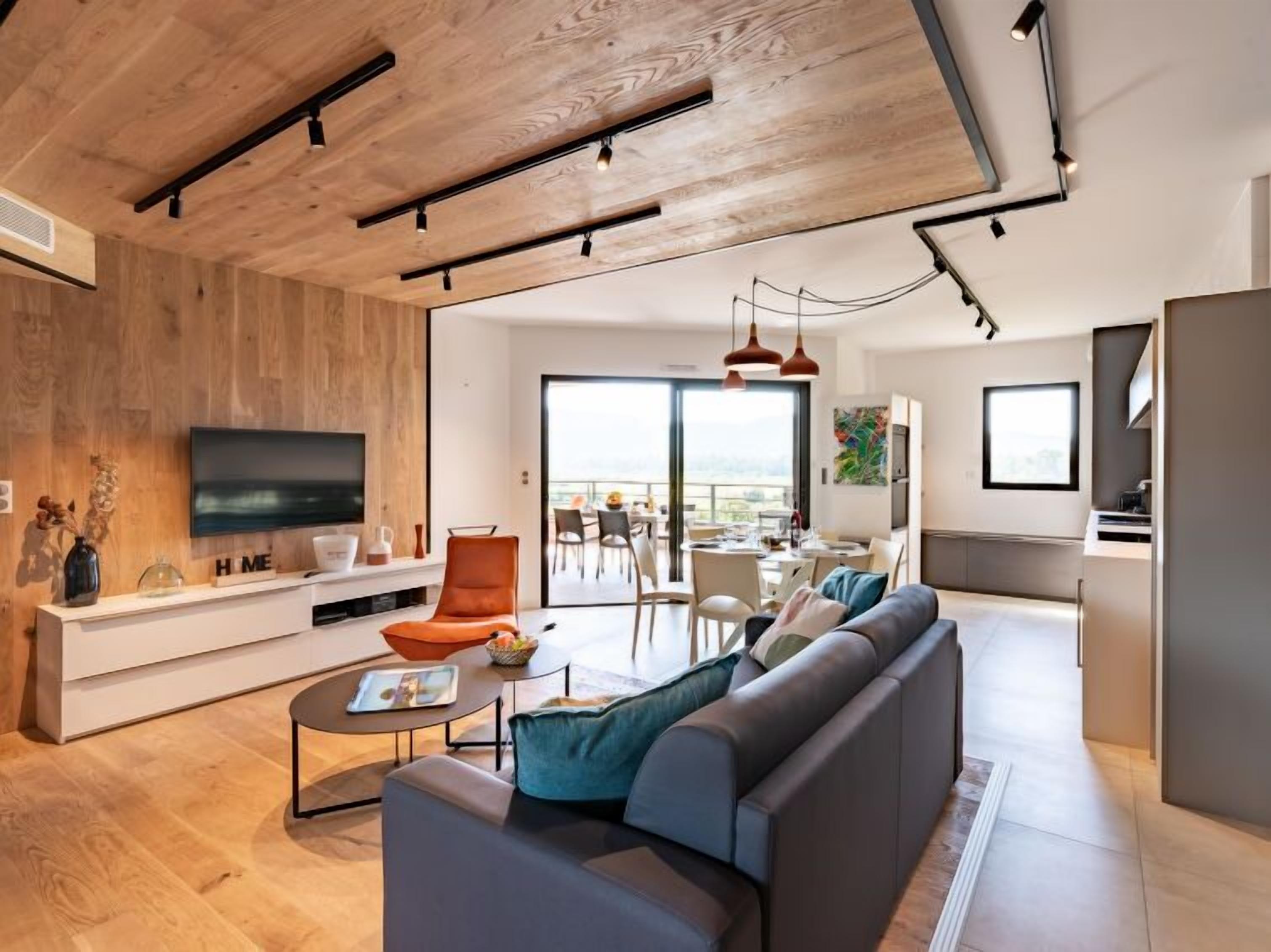 A modern living room with a wooden ceiling, a TV, a sofa, and a dining area with a view.