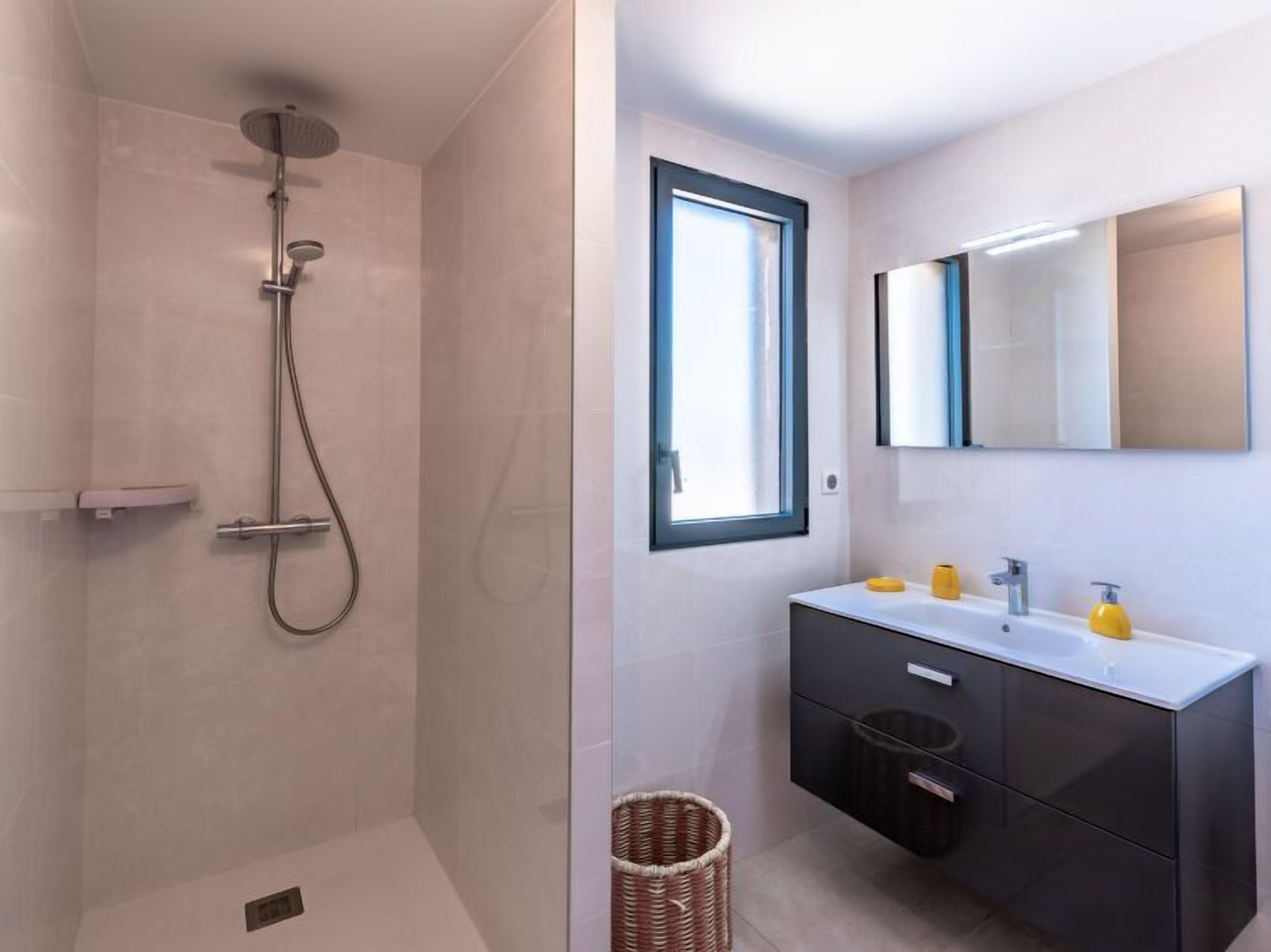 Un baño moderno con ducha envuelta en vidrio, un espejo grande sobre el lavamanos y un ventanal que proporciona luz natural.