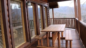 A wooden deck with a table and benches, large windows with blinds, and a view of mountains.
