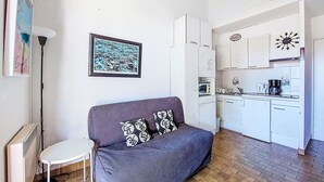A compact living space with a sofa, a small table, a kitchen area with white cabinets, and a broom on the ceiling. - Les Hauts du Port by Interhome (Saint Cyr sur mer Les Lecques)