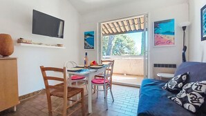 A cozy room with a bed, a dining table with chairs, a TV mounted on the wall, and a window with a view of trees and a beach. - Les Hauts du Port by Interhome (Saint Cyr sur mer Les Lecques)