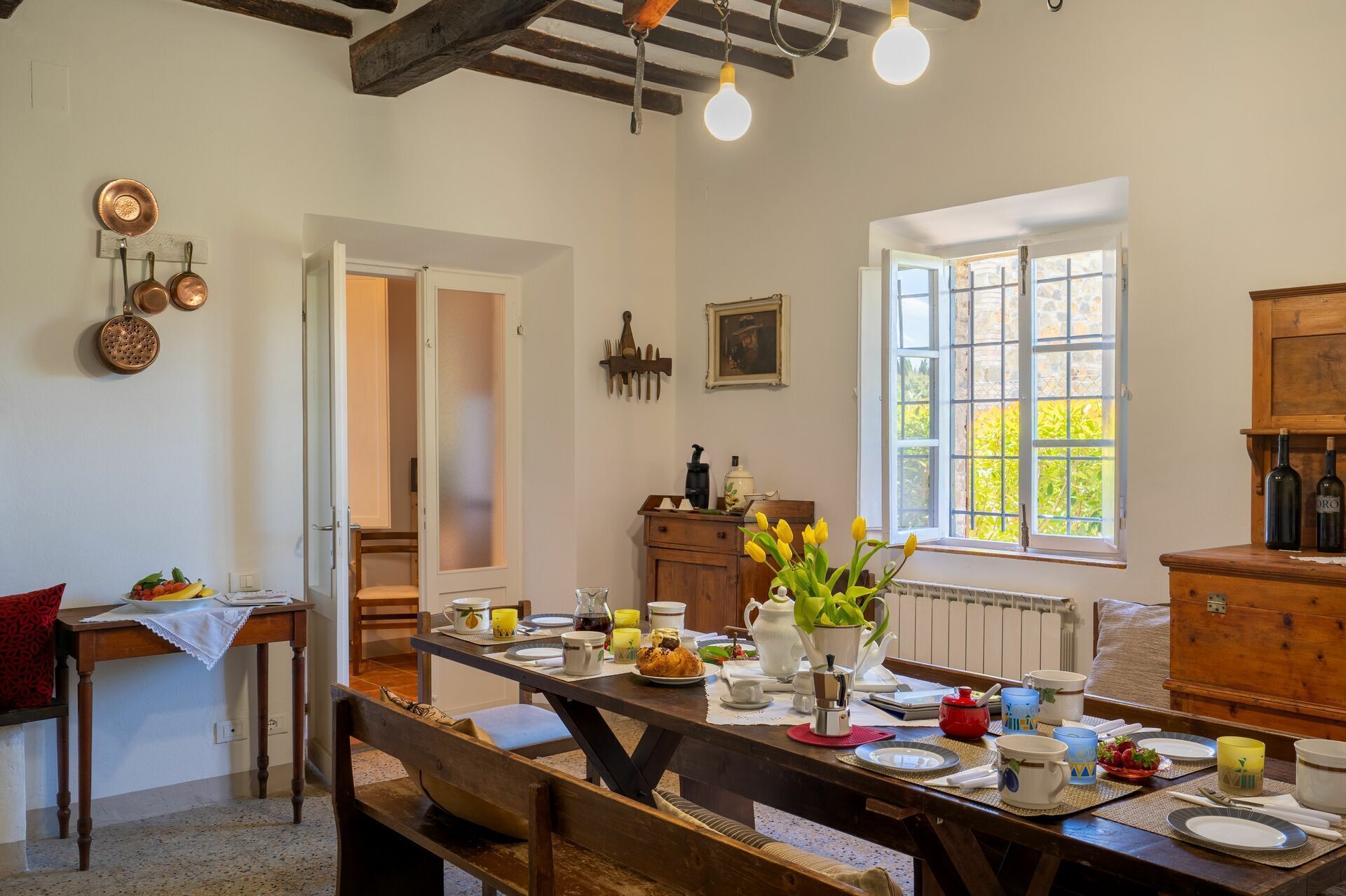 Un coin repas avec une table en bois dressée pour un repas, une fenêtre donnant sur un paysage verdoyant et un espace cuisine où l’on peut admirer des ustensiles de cuisine en cuivre accrochés au mur.