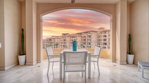 Varanda com mesa e cadeiras, um vaso de planta e vista para um conjunto de edifícios ao pôr do sol.
