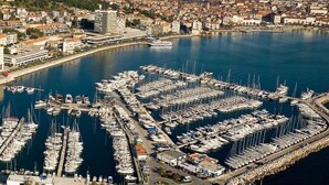 Vista aérea de uma marina com vários barcos atracados, cercada por uma paisagem costeira com edifícios e montanhas ao fundo.