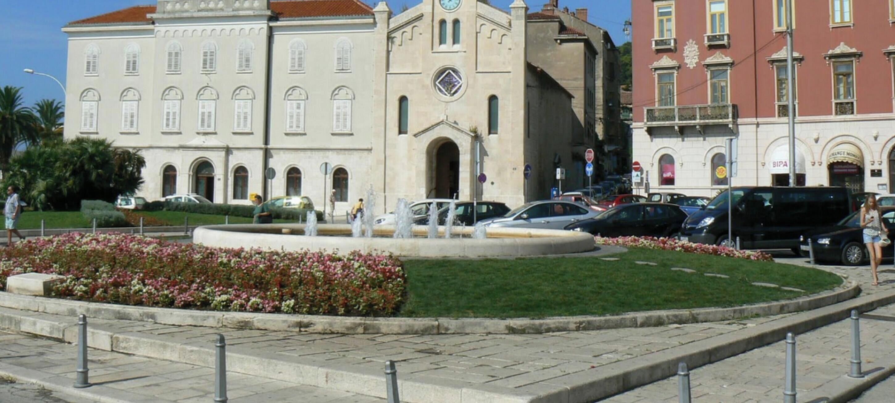 A public square with a fountain, surrounded by flower beds and a building with classical architecture.