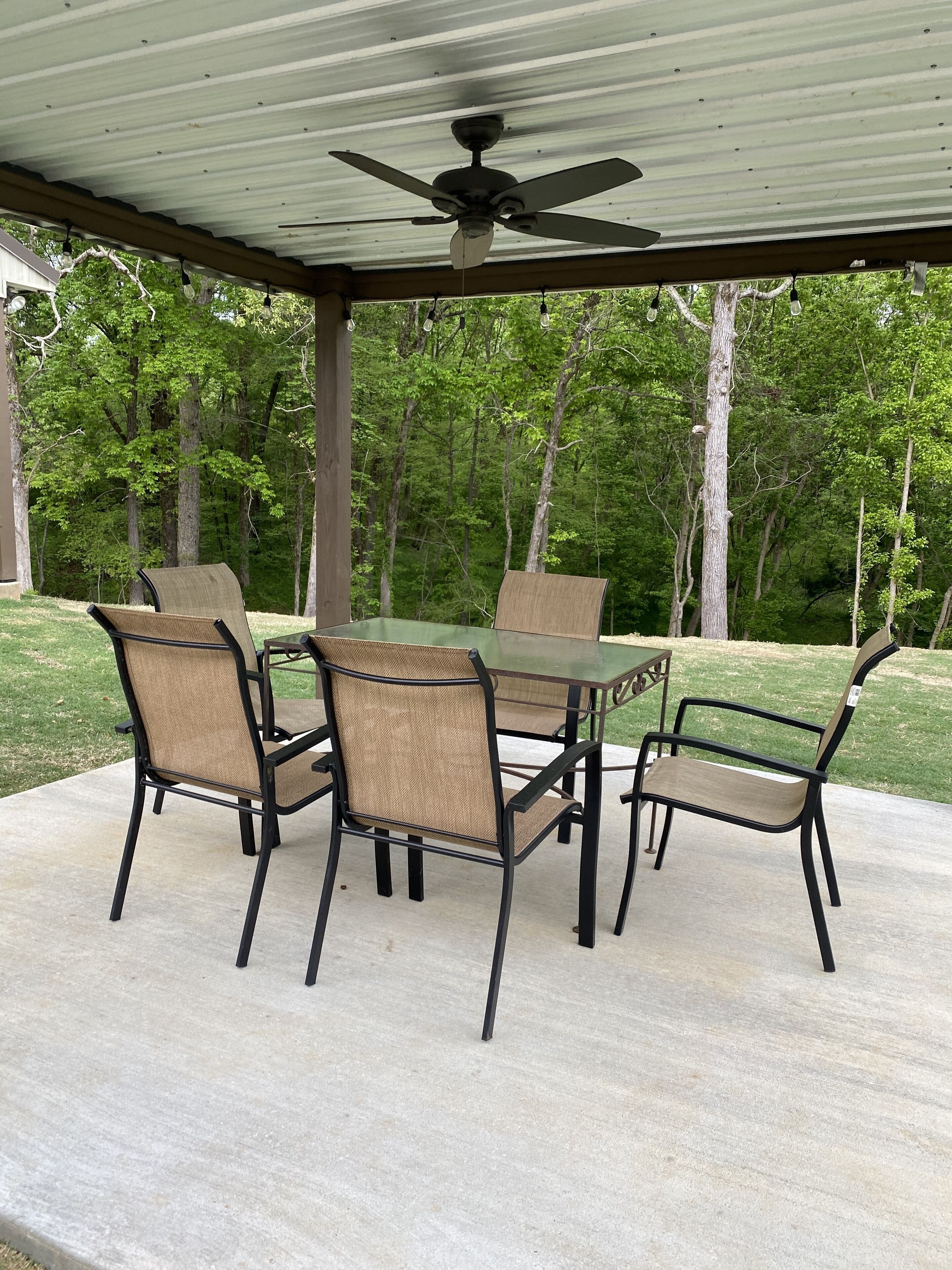Une terrasse extérieure avec une table et des chaises, un ventilateur de plafond et une vue sur une zone boisée.