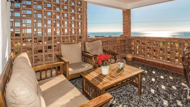 A balcony with wooden furniture, including a sofa and chairs, a small table with a vase of flowers, and a view of the sea.