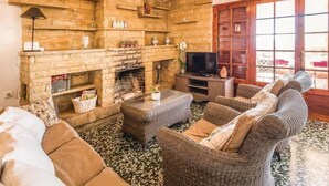 A cozy living room with a stone fireplace, wicker furniture, a television, and a large window with wooden shutters.