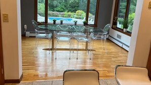 Un comedor con una mesa y sillas de vidrio, amplias ventanas con vistas a una piscina y follaje, y techo de madera.