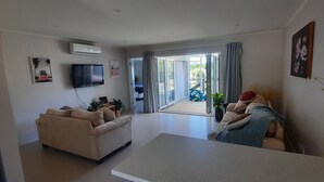 A living room with a sofa, a coffee table, a TV, and a sliding glass door leading to a balcony. - Walking distance to cafes and restaurants (Mangawhai)