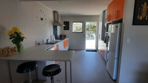 A modern kitchen with a white countertop, orange cabinets, a stainless steel refrigerator, and a black clock on the wall. - Walking distance to cafes and restaurants (Mangawhai)