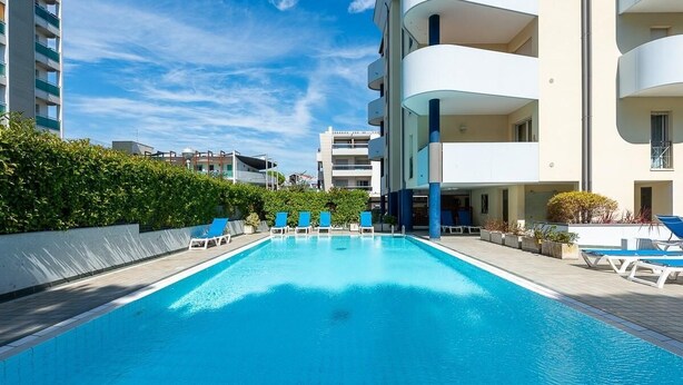 Una piscina con sillones de descanso, rodeada de follaje y edificios modernos.