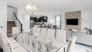 Una sala de estar moderna con una mesa de comedor de vidrio, sillas blancas, un sofá gris, un televisor montado en una pared de piedra y una escalera que sube a otro nivel.