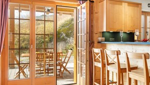 Een houten interieur met een schuifglazen deur die uitkomt op een terras, houten meubels en een keuken met een aanrecht en glazen.