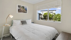 A bedroom with a bed, a bedside lamp, a window with a view of a house and greenery, and a framed picture on the wall.