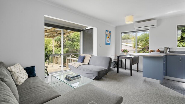 A modern living room with a grey sofa, a glass coffee table, and a dining area with a table and chairs. There is a kitchen area with a counter and cabinets, and large windows providing views of the outdoors.
