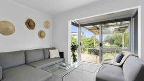 A modern living room with a grey sofa, a glass coffee table, and a grey armchair. There are three hats on the wall and a sliding glass door leading to an outdoor patio.