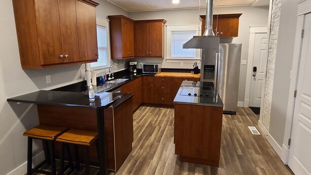 A modern kitchen with wooden cabinets, a central island, bar stools, stainless steel appliances, and a window with blinds.