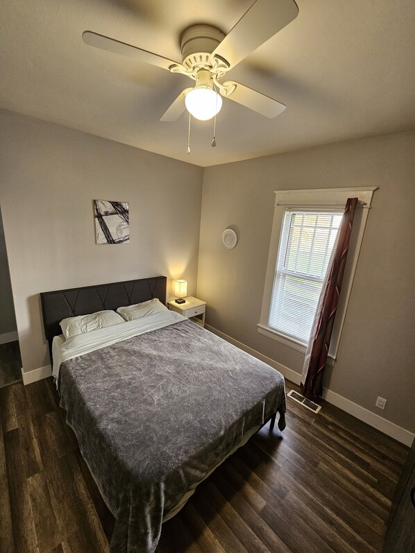 Un dormitorio con cama, ventilador de techo, ventana con cortinas, lámpara de noche y un reloj de pared.