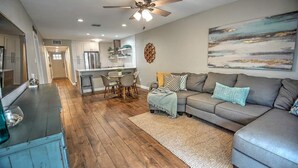 A modern living room with a grey sofa, wooden flooring, a ceiling fan, and a kitchen area in the background.