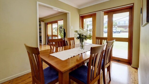 A dining area with a wooden table and chairs, a vase with flowers, and large windows with a view of the outside. - Retreat on Gleeson (Forster)