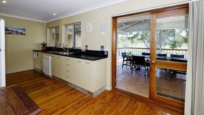 A kitchen with wooden flooring, a dining area with a table and chairs, and a sliding glass door leading to an outdoor patio. - Retreat on Gleeson (Forster)