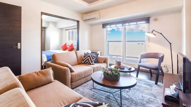 A modern living room with a sofa, armchair, and coffee table. There is a TV, a lamp, and a window with a view. - Glow (Otaru)