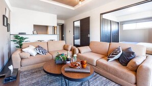 A modern living room with a beige sofa, a coffee table, and a dining table. There is a plant on the left side and a window on the right side. - Glow (Otaru)