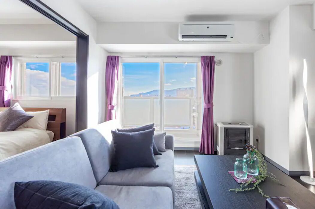 A modern hotel room with a large window, a sofa, a table with decorative items, and an air conditioning unit.
