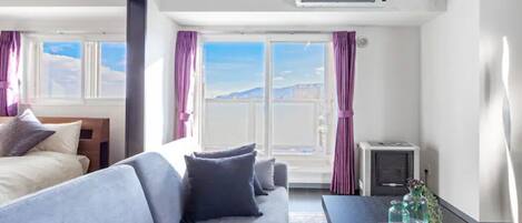 A modern hotel room with a large window, a sofa, a table with decorative items, and an air conditioning unit.