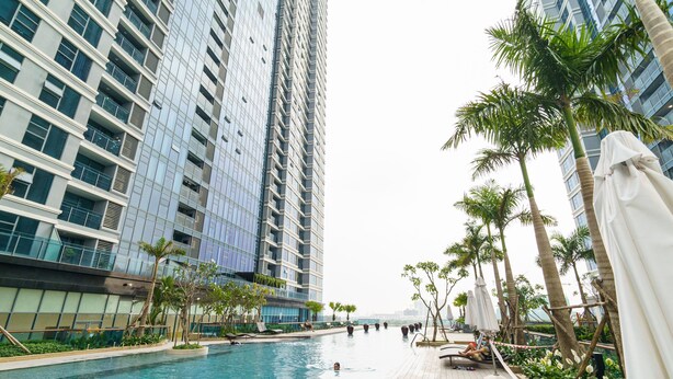 A swimming pool area with a wooden deck, surrounded by tall buildings and palm trees. - Hoasun Skyview Sunwah Pearl (Ho Chi Minh City)