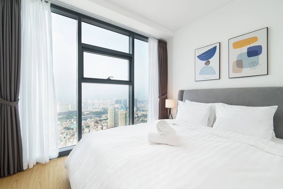 A hotel room with a large window, a neatly made bed, and two abstract art pieces on the wall.