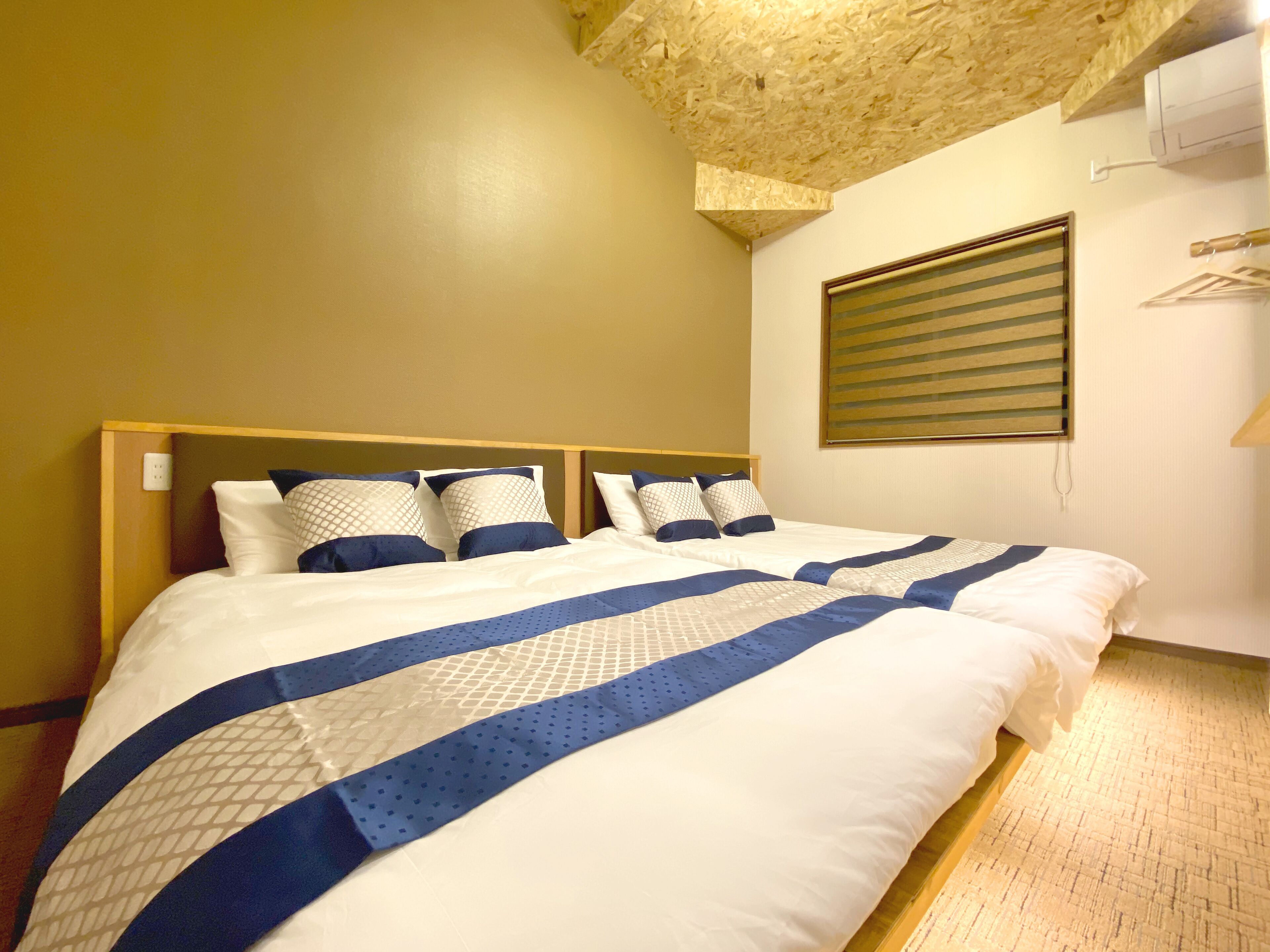 A neatly made bed with a blue and white striped bedspread and matching pillows, a wooden headboard, and a window with blinds.