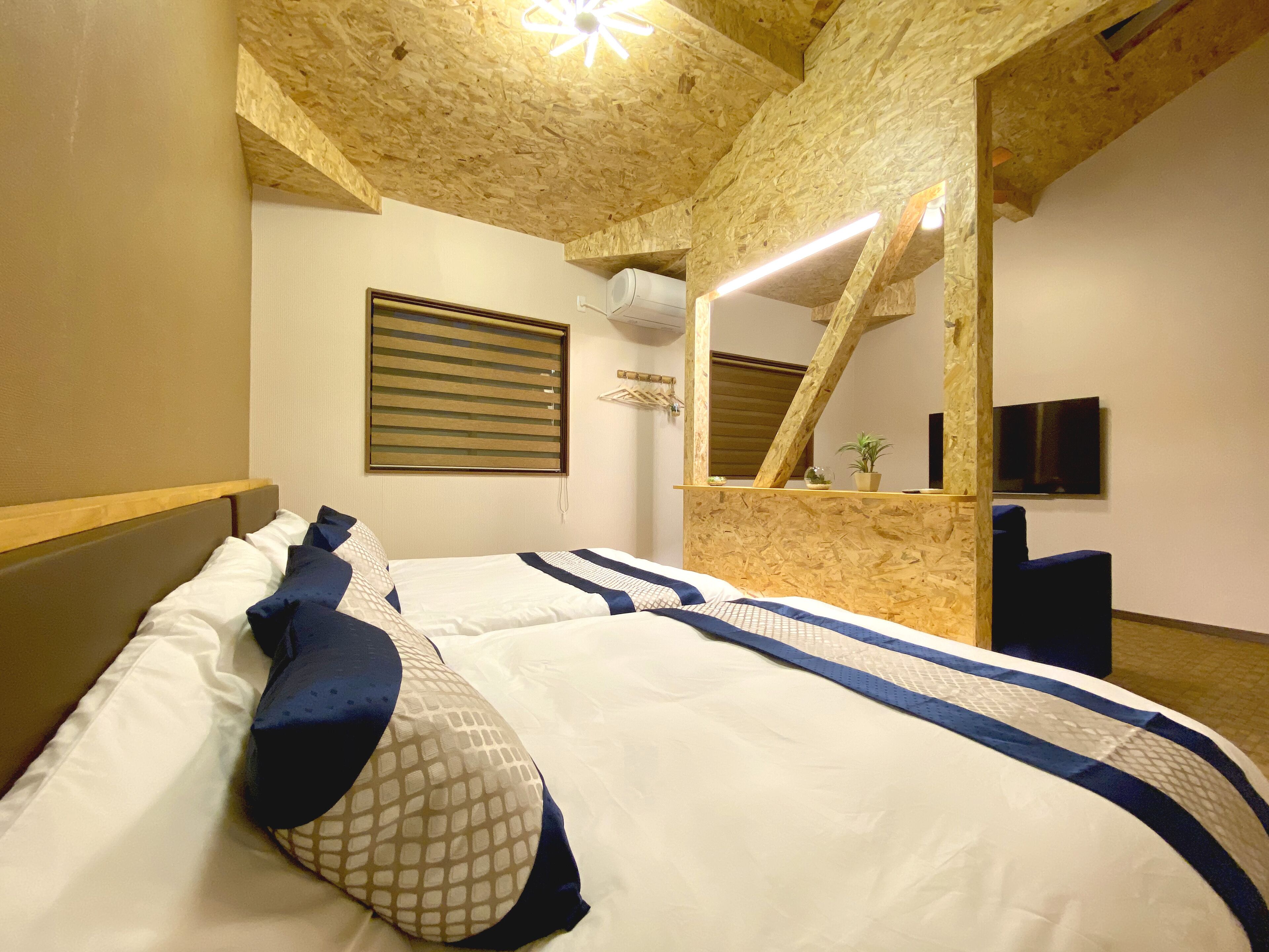 Un dormitorio con techo de madera, cama con ropa de cama blanca y azul, televisor montado y una planta pequeña en un estante.