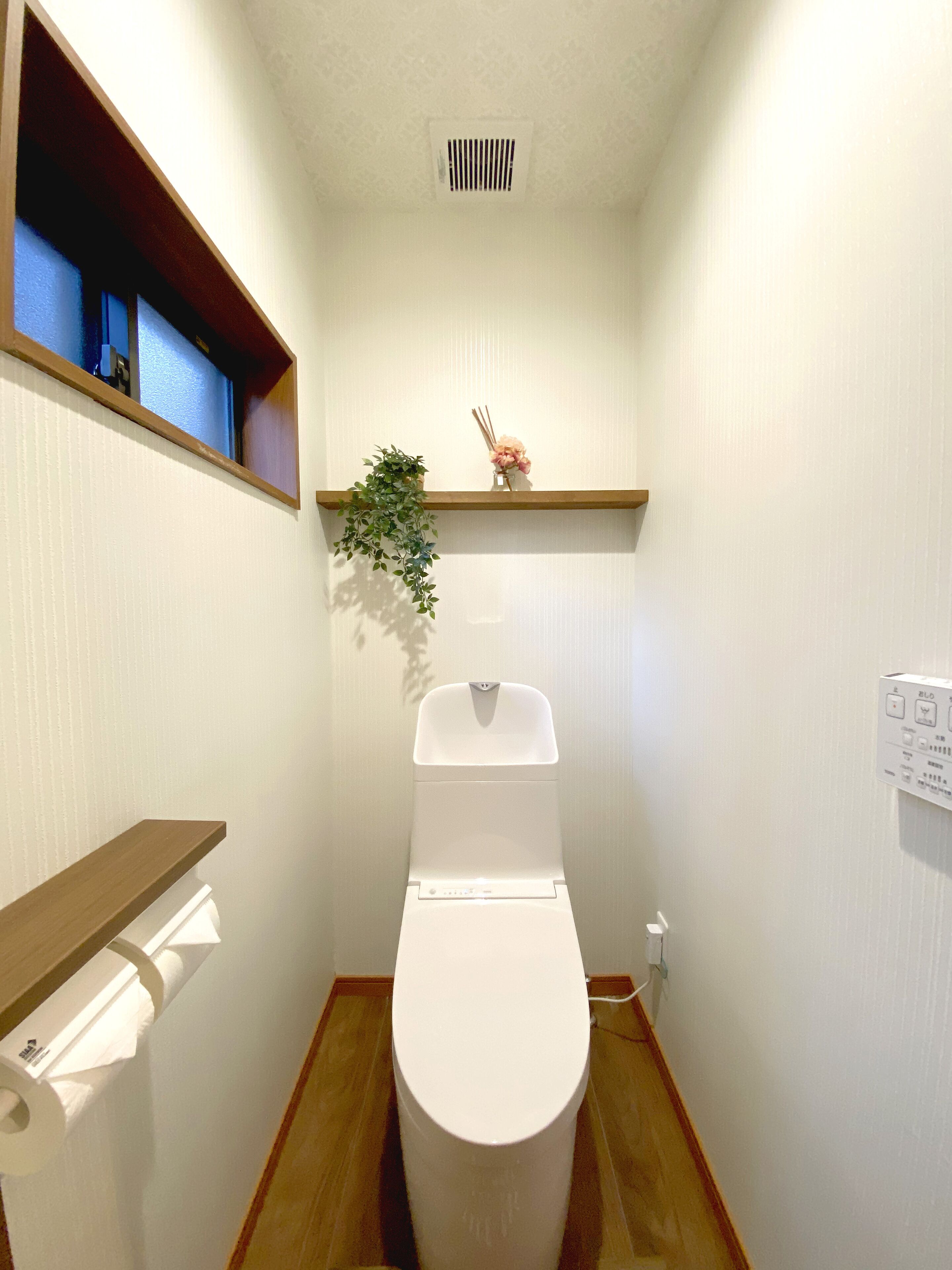 A small, compact bathroom with a toilet, a wooden shelf, a small plant, and a wall-mounted electrical outlet.