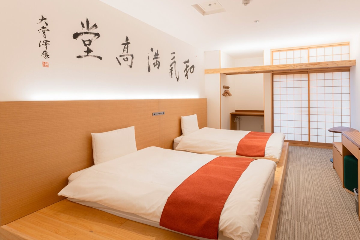 Habitación de hotel con dos camas, un escritorio, una puerta corredera tradicional y caligrafía japonesa en la pared.